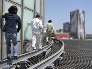 開放感のある場所。ビルの上の写真。一本橋のような細い足場を3人の人が歩いている様子。