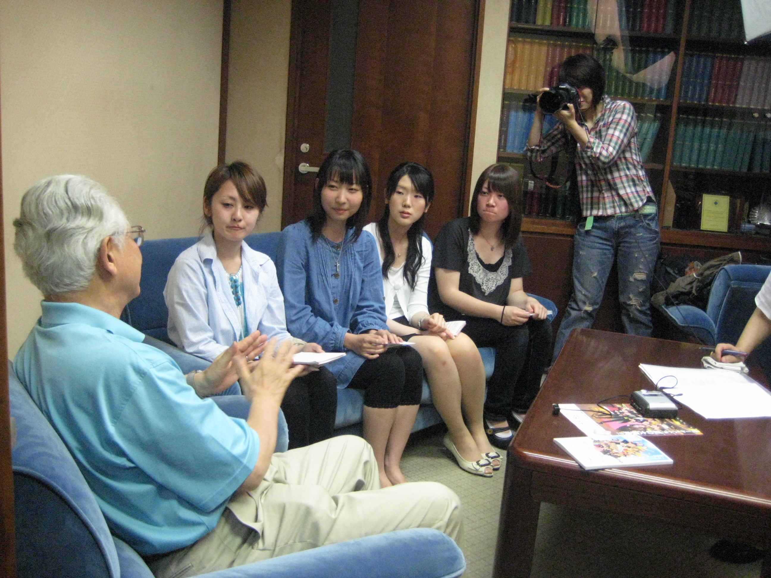 左手前に秋山さん、近くのソファーに4人の学生が座っている。
