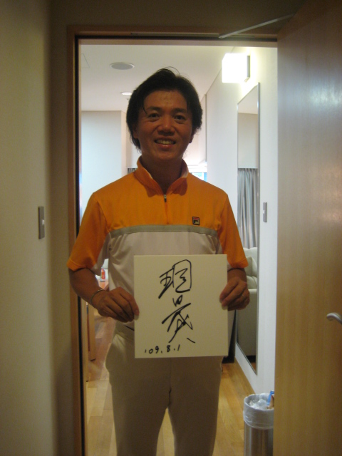 オレンジの服を来た現田マエストロの写真、。自身のサインが書かれたサインボードを両手にもっている。笑顔。