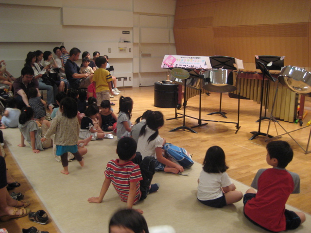楽器が並んだステージの前に絨毯が敷かれていて、子どもたちが座っている。