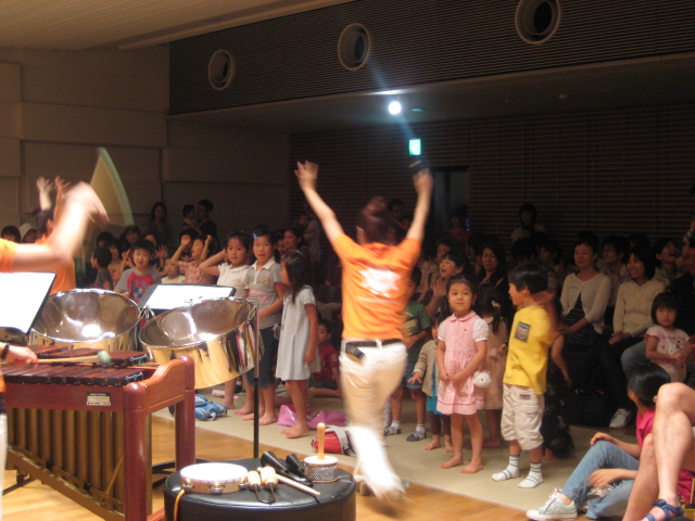 出演者が万歳をしながら子どもたちに呼びかけている様子。子どもたちは立ってステージを向いている。 width=