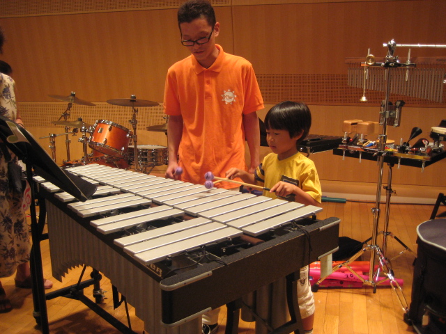 オレンジの服を着た出演者の人に教わりながらヴィブラフォンを体験している黄色い服の子どもの写真。