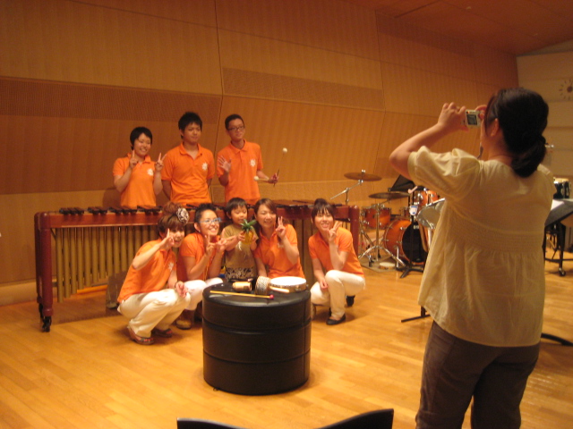 出演者の記念写真を撮っている様子を写した写真。8人の出演者はオレンジの服を着ていて、中央に熱狂的ファンならぬ子どもがいる。