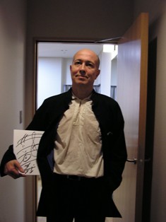 白いシャツに黒いジャケットを来た井上マエストロの写真。右手に直筆サインが入ったサインボードを持っている。きりっとした顔。