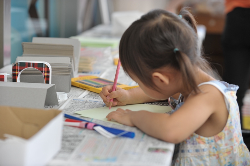 柔らかそうな髪の毛を水色の髪留めでツインテールに結んだ小さな子どもが、ピンク色の色鉛筆を右手に持ち、紙に色を塗っている様子。机の上には汚れ防止用の新聞紙が敷いてある。