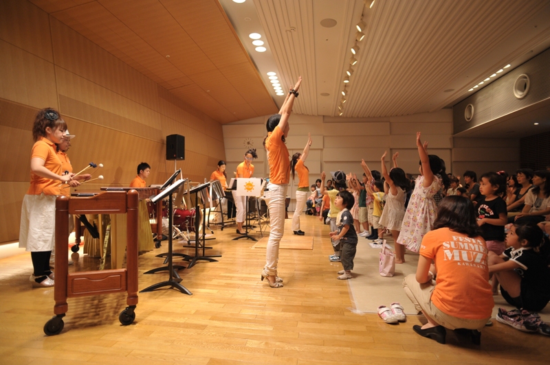 市民交流室の下手横からステージを写した写真。マリンバや出演者、子どもたちが写っていて、楽しそうに両手を上げて音楽を楽しんでいる様子。