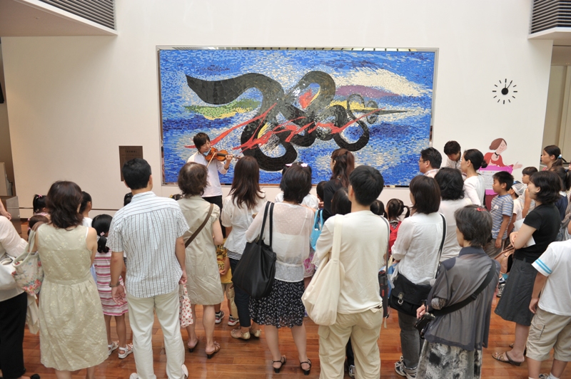 ホール2階ホワイエ、岡本太郎の美術品の前で高木さんが演奏しているのを立って聴いている大人や子どもたちの後ろ姿が写っている。