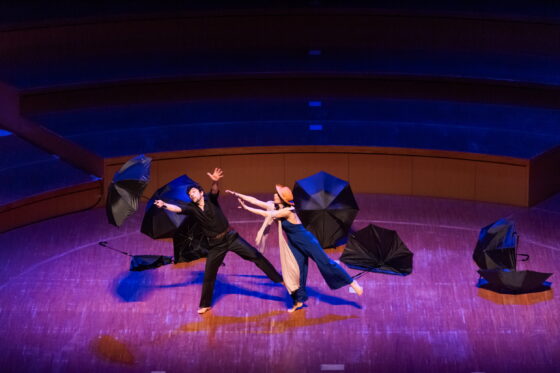 薄暗い舞台の中央で、傘を持ちとばされそうな男性（左）と、そこに手を差し伸べる女性（右）