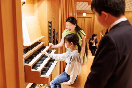 演奏台（メインコンソール）で左右から龍田さんと丁さんに見守られながら鍵盤に指を乗せる子ども