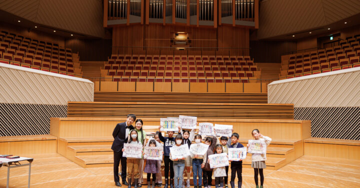 ステージ中央で、完成した塗り絵を体の前や頭の上で見せる12名の子どもたちの左側に龍田さんと丁さんの集合写真