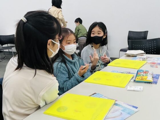 隣り合う人と顔を見合わせ、胸の前で手を開き身振りも加えながら話し合う3人のジュニア・プロデューサーたち