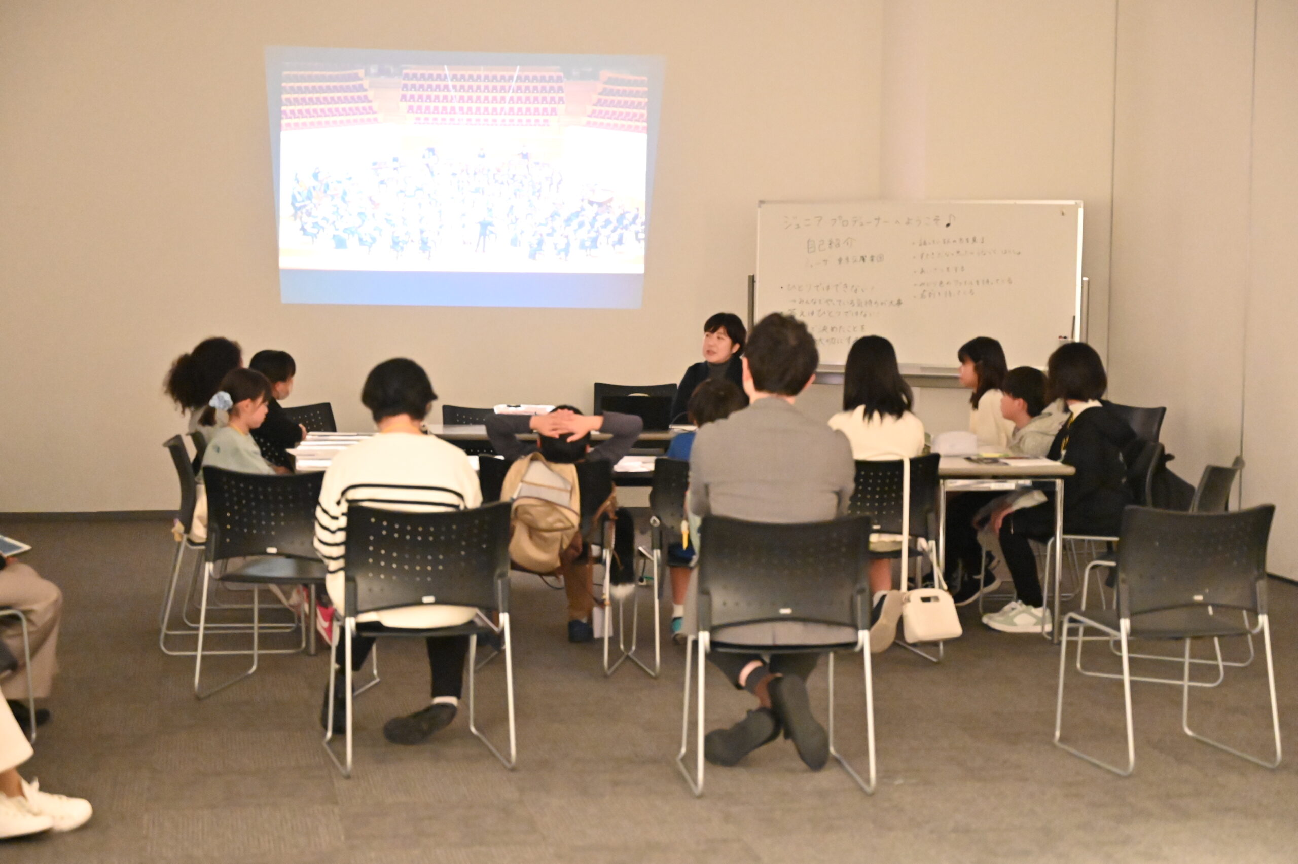 前方に東京交響楽団のスタッフ桐原さんが座り、周りで着席しプロジェクターを見ながら話を聞くジュニア・プロデューサーたちとスタッフ