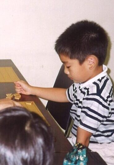 将棋大会で将棋を指す幼少期の熊倉さん。真剣な表情で盤面を見ている。