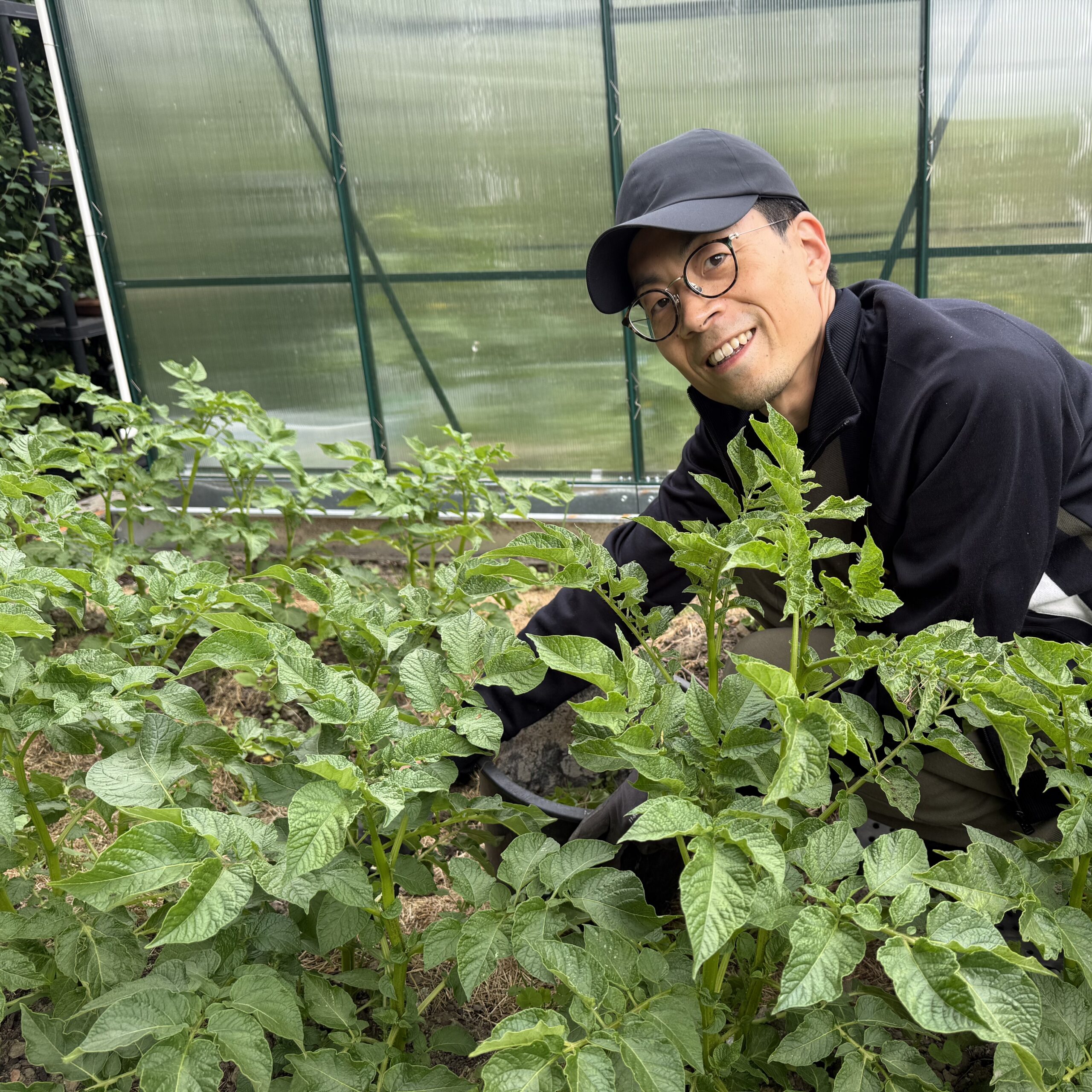 育てている作物のわきにしゃがみ、作物越しにカメラの方を向く小林さん。