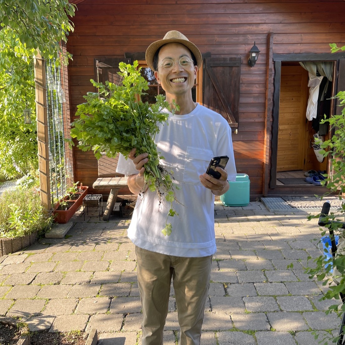 収穫物を持ち笑顔で写真に写る小林さん。Tシャツにハット、チノパンのとてもラフな服装。