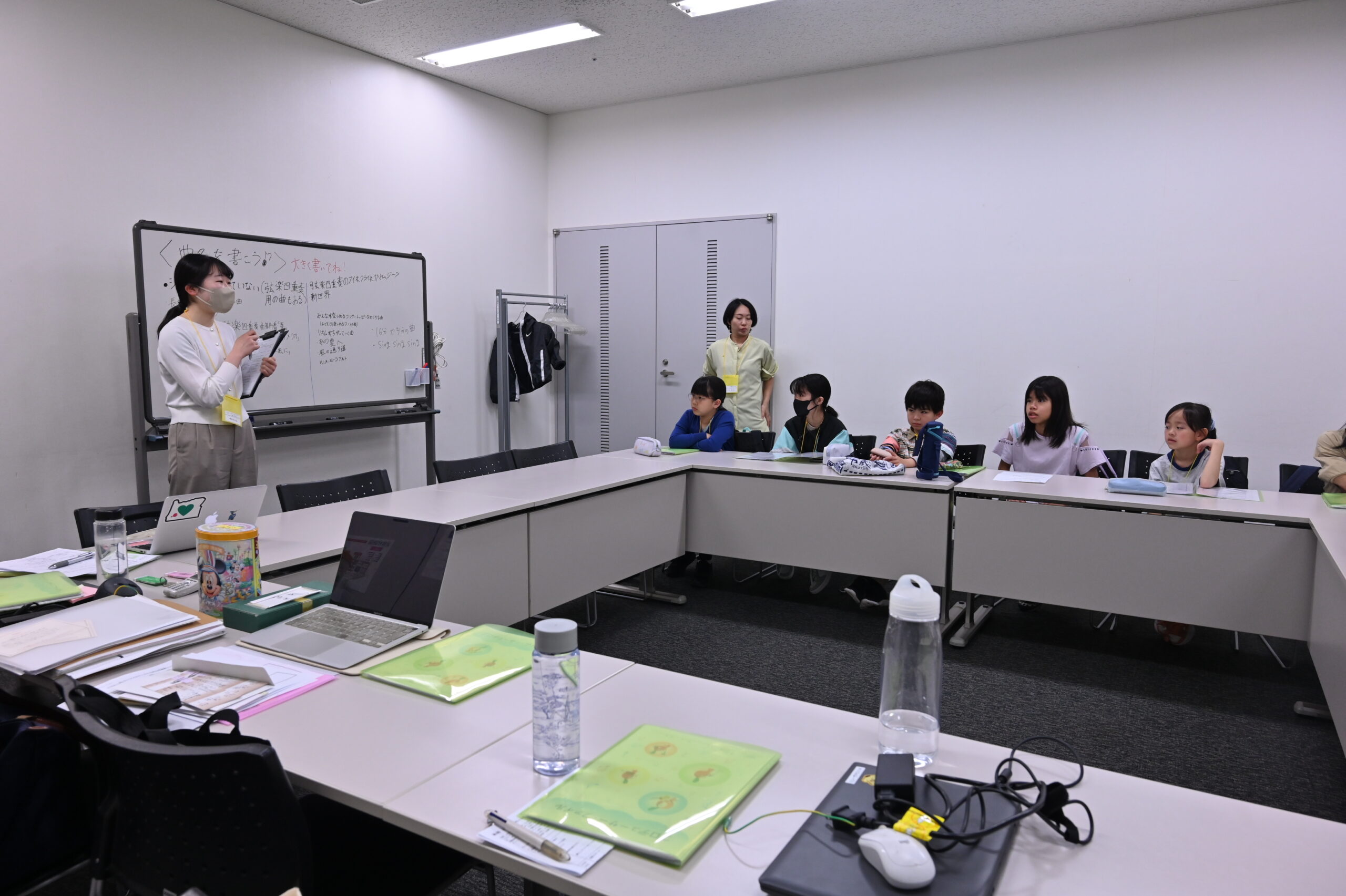 最初に全員で集まって話を聞いている様子の写真。ロの字に並べられた机の2辺にメンバーが座り、ホワイトボードの前に立つスタッフの方を見ている。