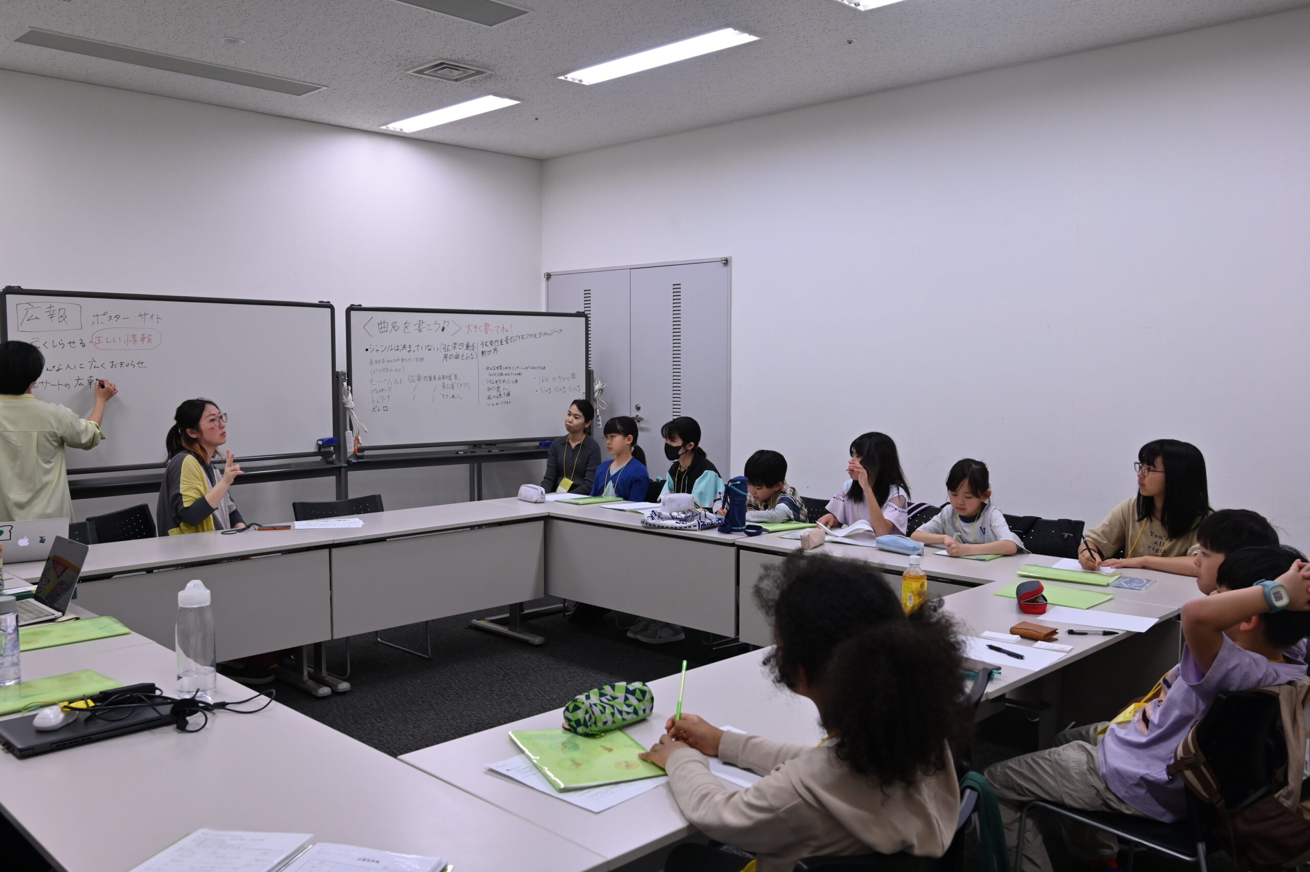 広報スタッフの森さんから話を聞くジュニア・プロデューサーたちの写真。メンバーはメモを取ったり、森さんの方を見ていたりしている。ホワイトボードの前に森さんが着席し、書記スタッフもいる。