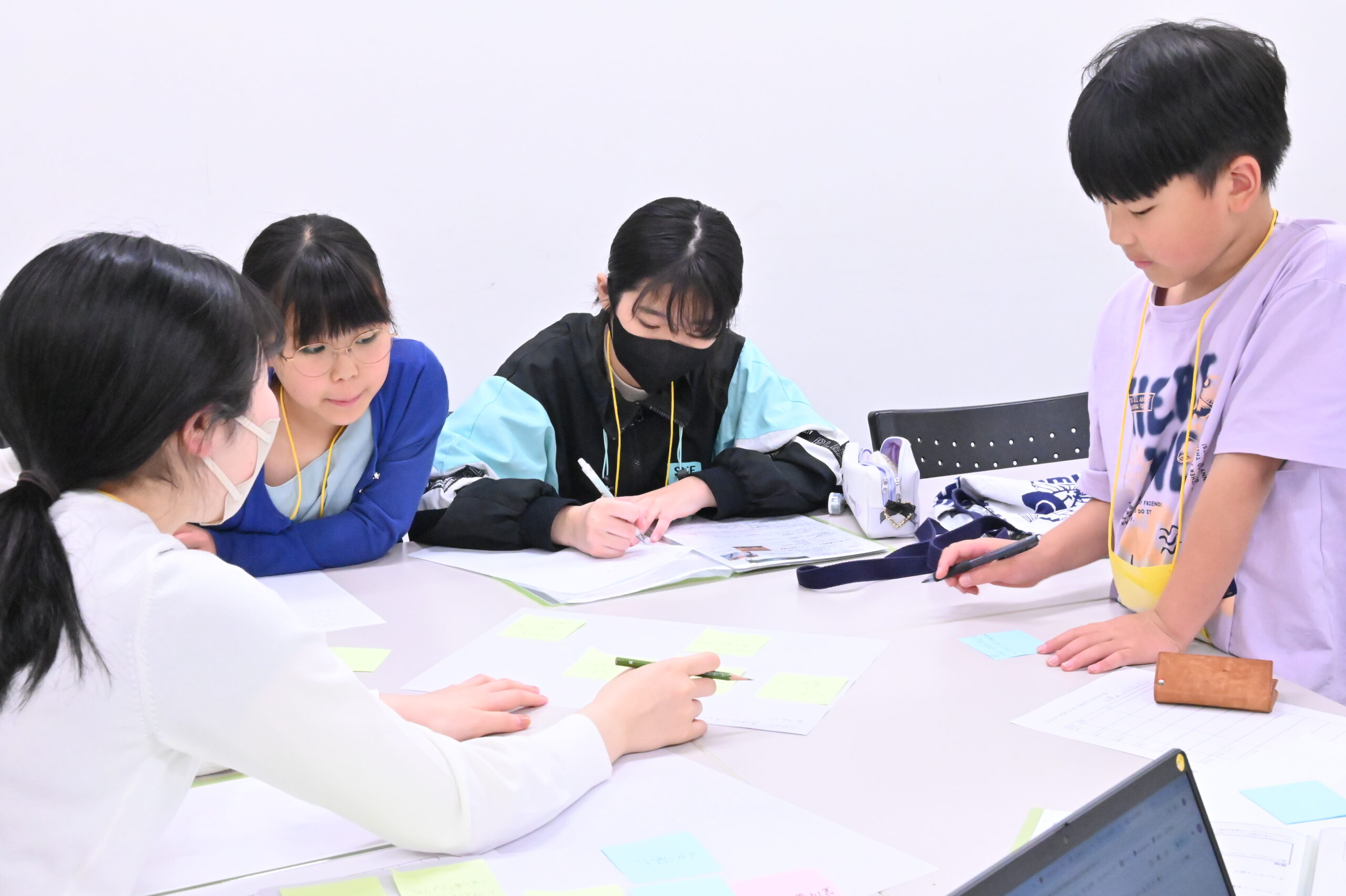 話し合い中の企画班の写真。メンバー2名とスタッフは着席し、1名は立ちながら、机の中央に置かれた付箋つきの紙を見ている。
