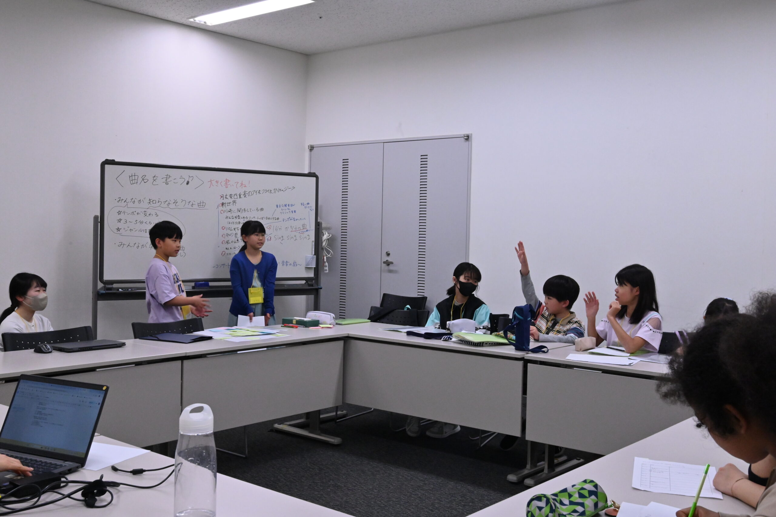 今日決まったことを全員に共有しているところの写真。メンバー2人がホワイトボードの前に立ち、他のメンバーは挙手したり、周囲を見たり、メモしたりしている。