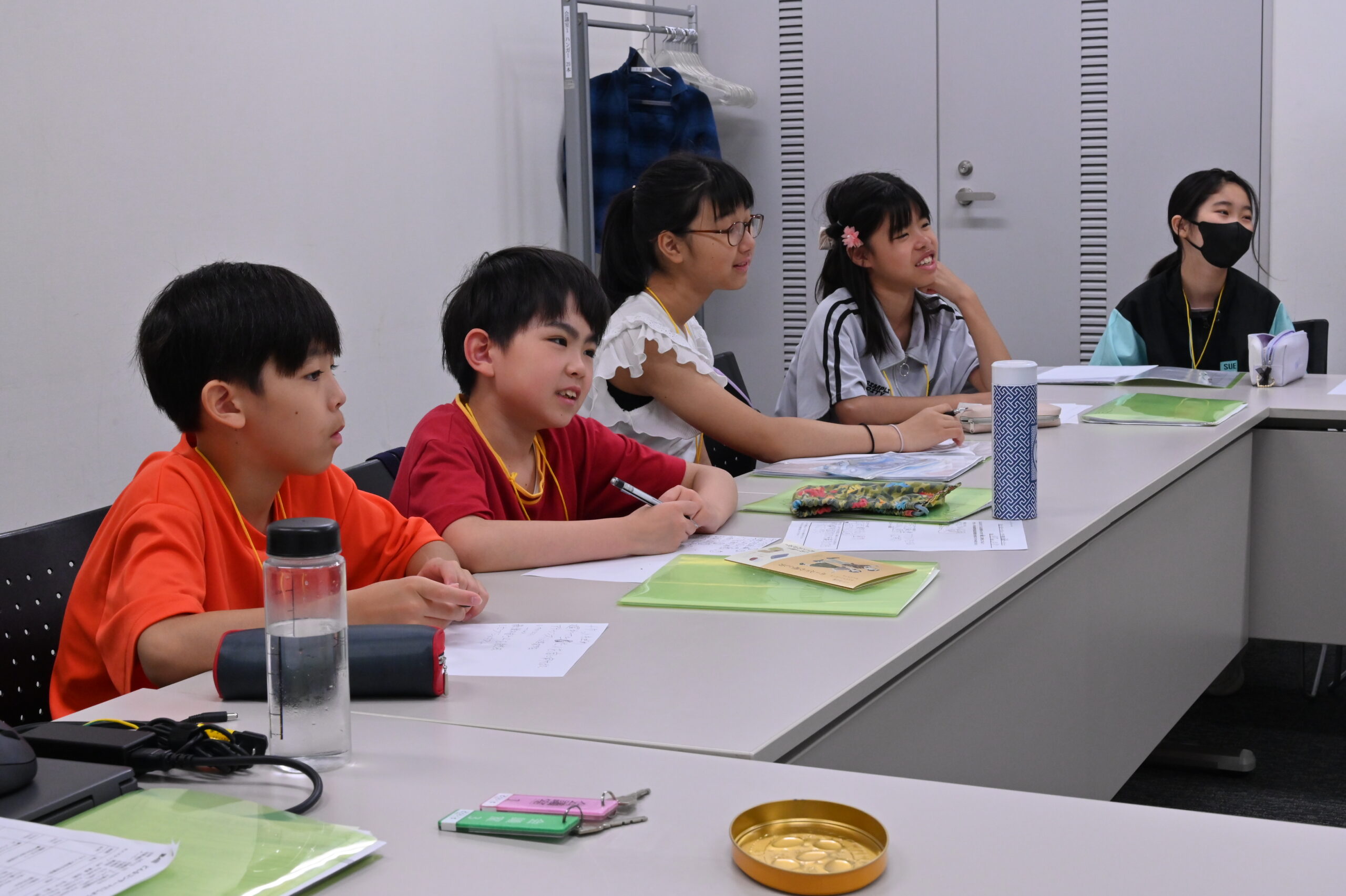 並んで座り同じ方向を向いて、各々何かを考えていたり、にこやかに話に参加している様子のジュニア・プロデューサー5人の写真。手元には筆記用具やファイル、水筒が置かれている。