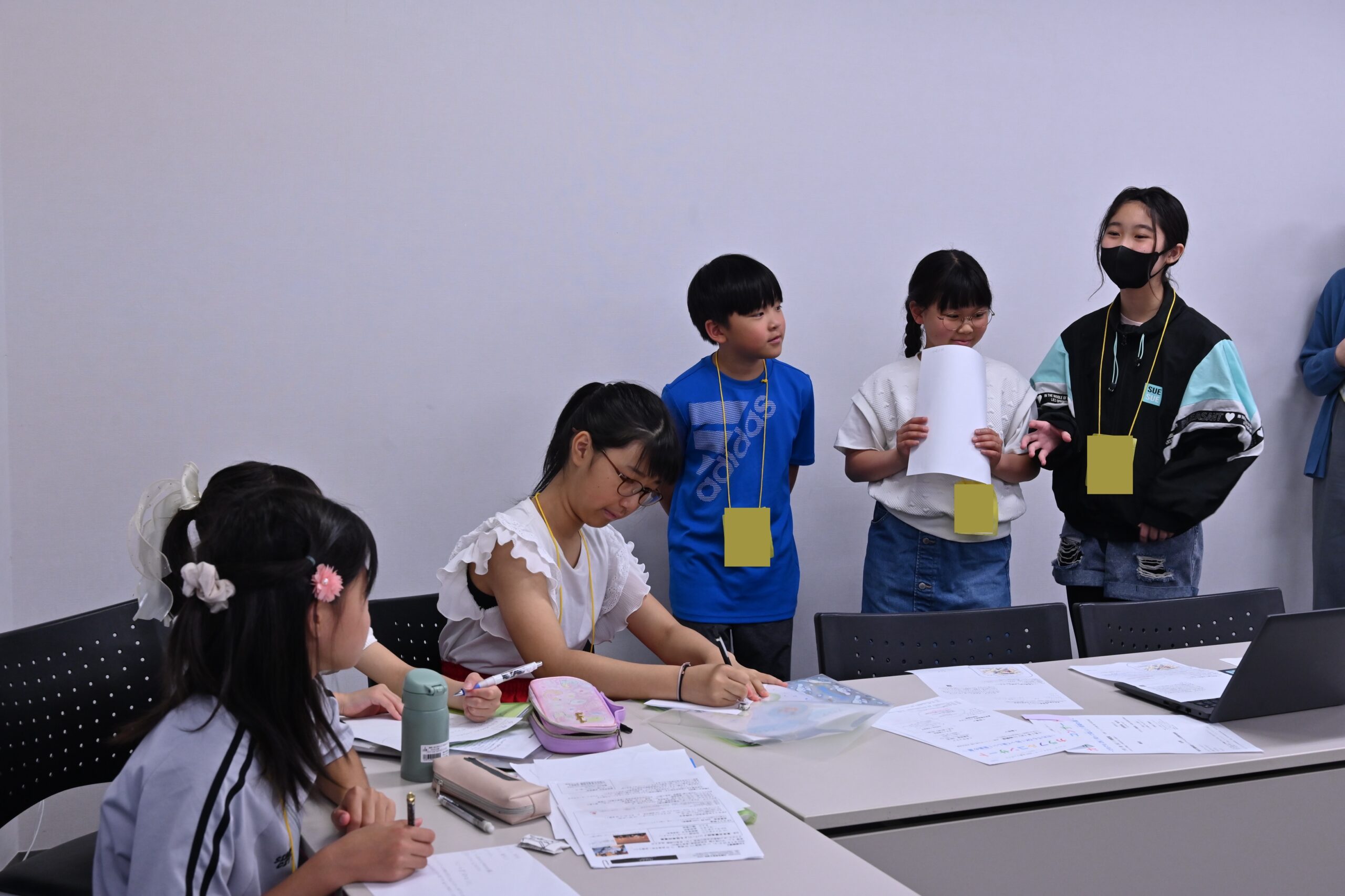 写真の右側には企画班3人が立って、左側の広報班に何かを伝えている様子。広報班メンバーは企画班の方を見たり、メモをしたりしている。