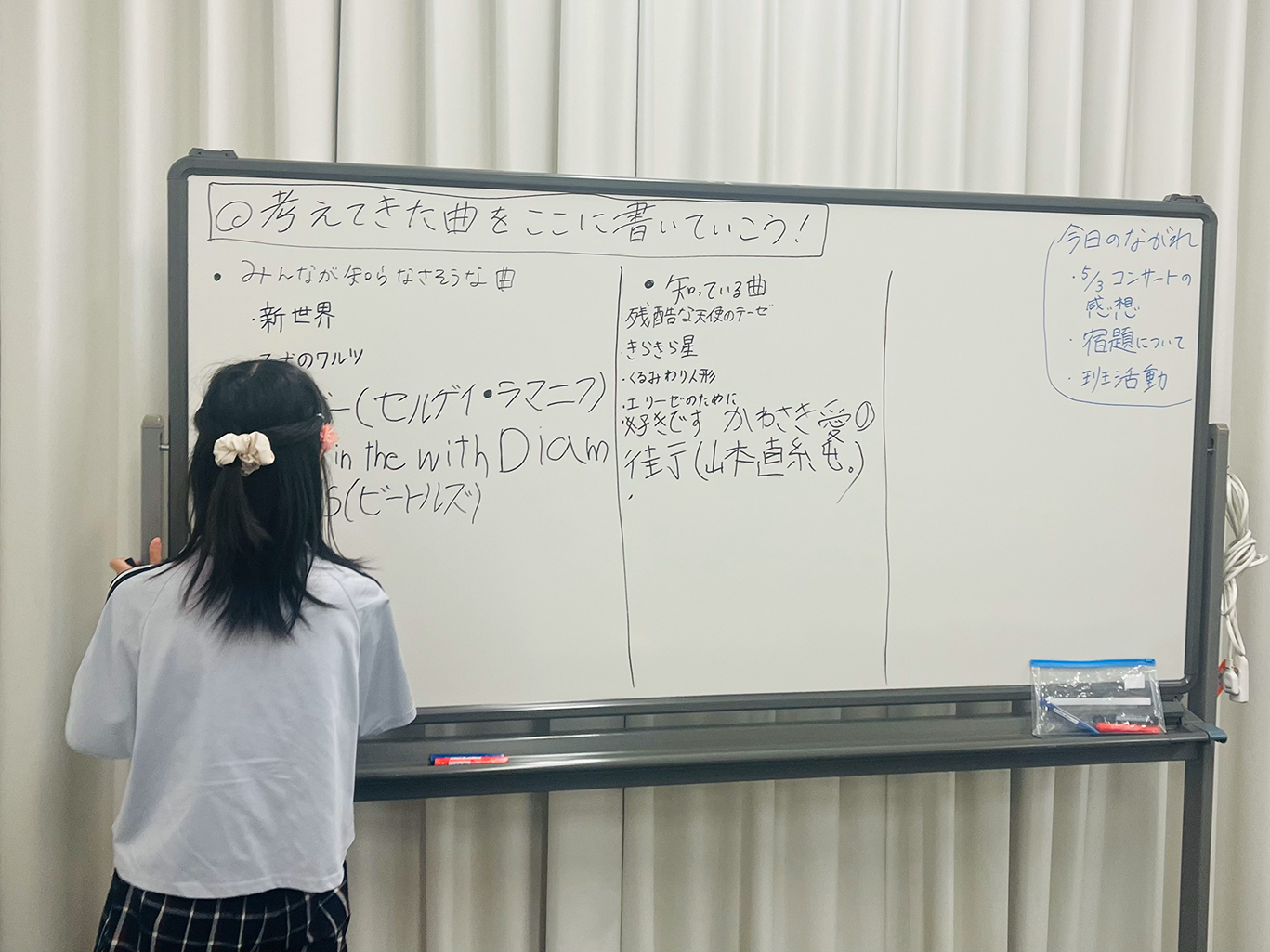 ホワイトボードに向かっている1人のジュニア・プロデューサーの後ろ姿の写真。「考えてきた曲をここに書いていこう」と書かれたホワイトボードには、「みんなが知らなそうな曲」に「新世界」「子犬のワルツ」等、「知っている曲」には「残酷な天使のテーゼ」「きらきら星」等5曲程ずつ既に書かれている。