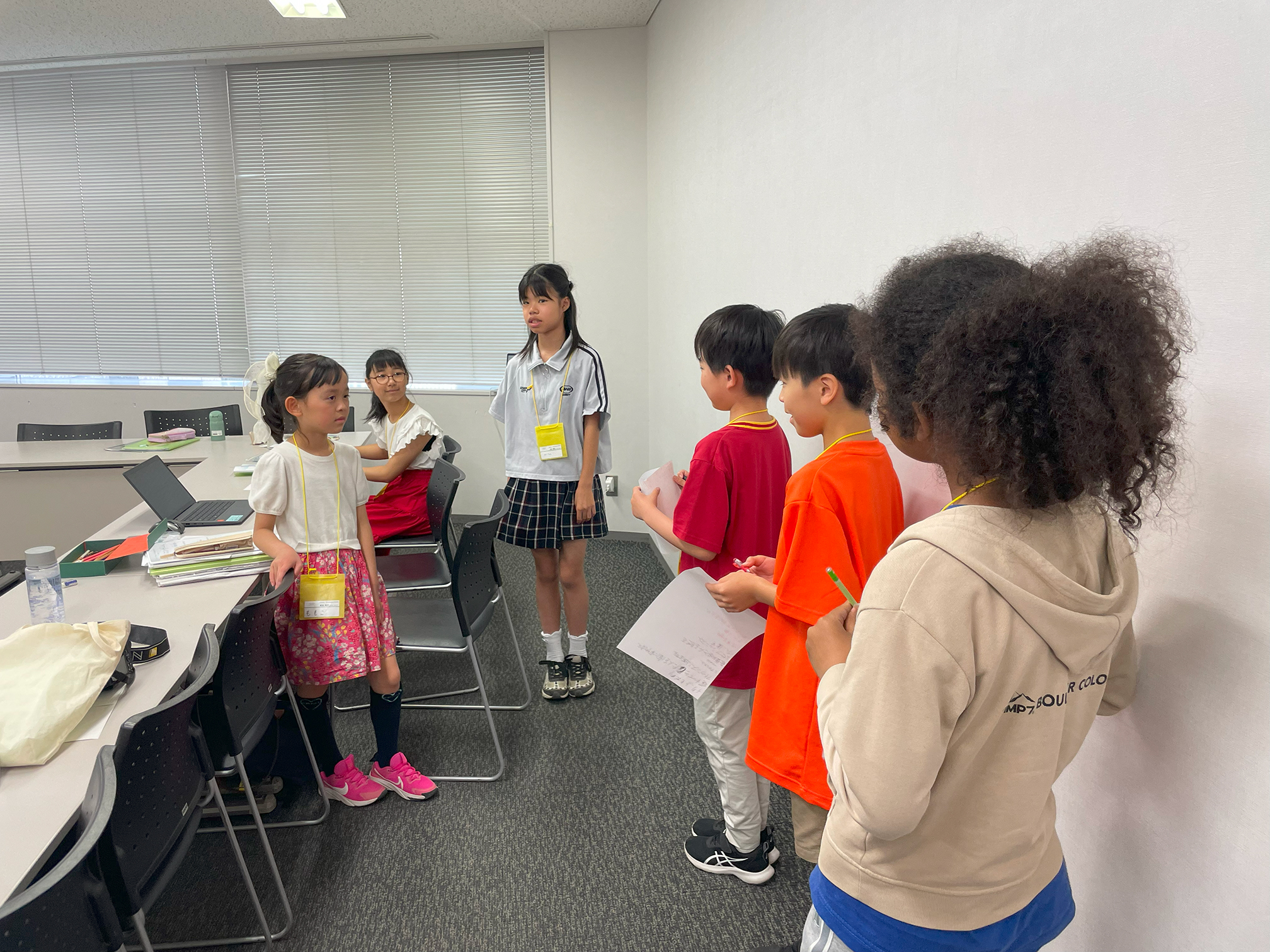 机の近くに立ったり座ったりしている広報班3人の元に近づき、壁側に1列に並ぶ運営班3人の後ろ姿の写真。