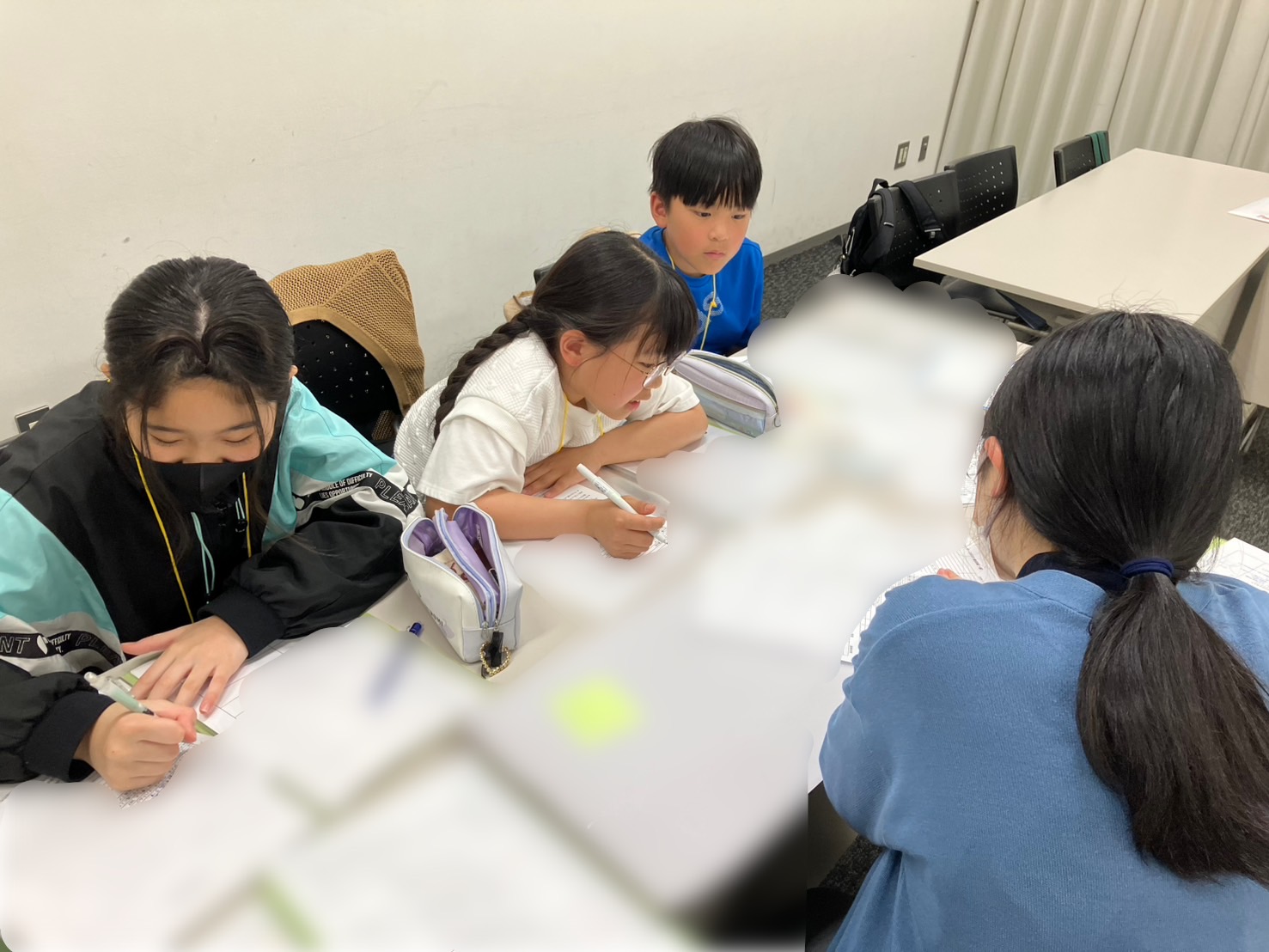 企画班3人横並びで座り、資料を見たりメモをしたりしている。3人の前にいる大人スタッフのゆみちゃんが資料を見せている。机上はモザイク処理されているが、沢山の資料が広げられている。