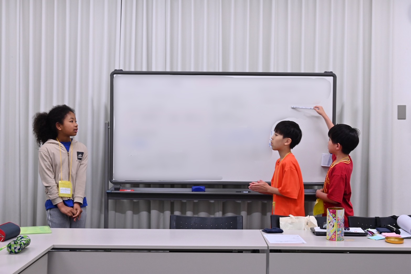 ホワイトボードを見ながら報告する運営班3人の写真。1人はホワイトボードの左側に立って話し、1人はホワイトボードの前に立ち話す子の方を見ており、後の1人はホワイトボードの右側に立ち、細長いマグネットのようなものでホワイトボードを指し示している。ホワイトボードはモザイク処理されている。