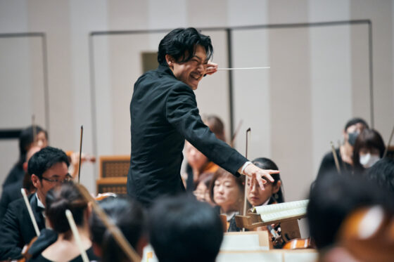 指揮棒を高く構え、険しい表情でオーケストラを見る出口さん。右手は大きく開いて力がみなぎっている。