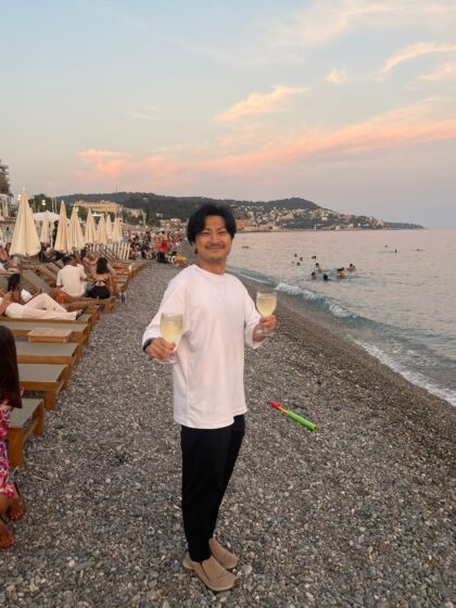 ワイングラスを両手に持ち、浜辺に立って笑顔でこちらを向く出口さん。