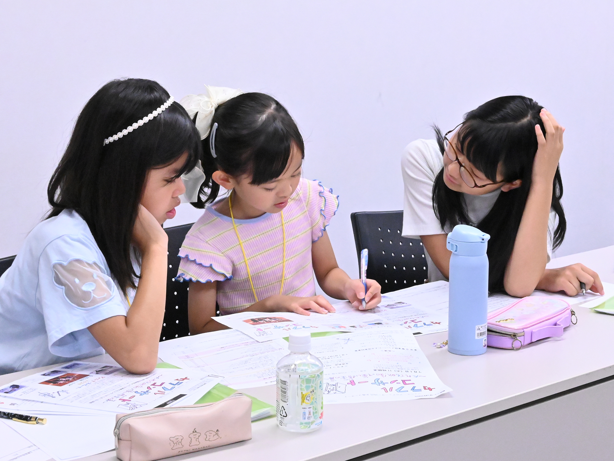 広報班3人が横並びに座り、1枚のプログラムを確認して、1人が何かを書き入れている写真。他にも机上には何枚かのプログラムやチラシが広げられている。