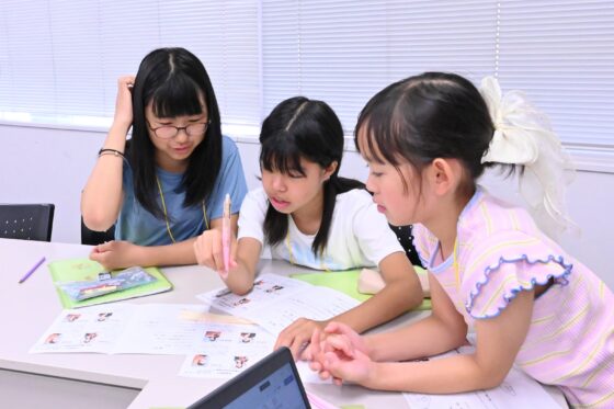 広報班メンバー3人が顔を寄せ合って、机上に並んだプログラムを確認している写真。