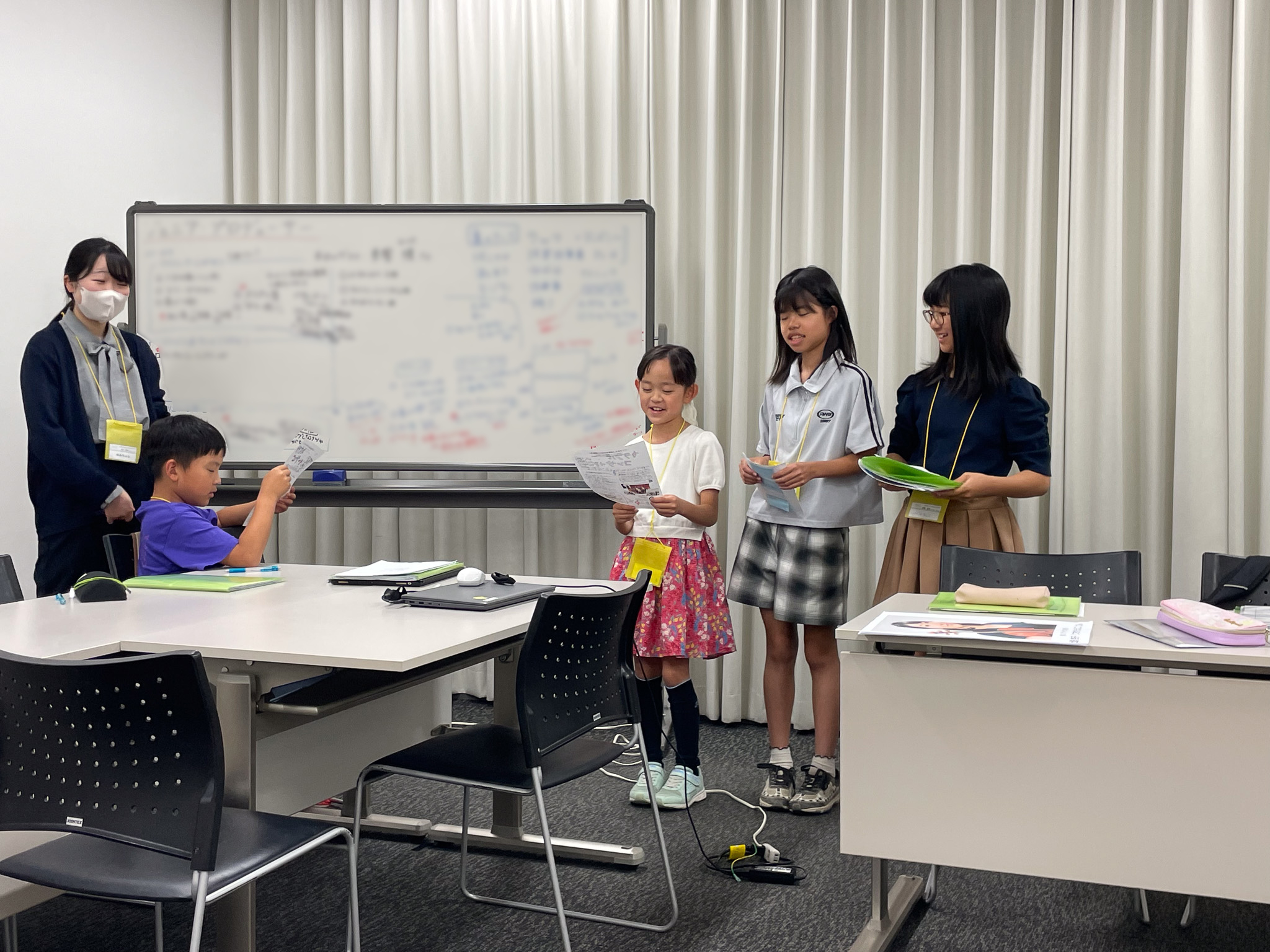 ホワイトボードの左横には大人スタッフ、右横に広報班3人が立って報告している様子の写真。手にはそれぞれ資料やファイルを持っている。