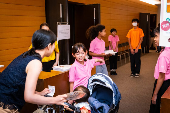 入口でベビーカーでお子様をお連れのお客様を笑顔で案内するジュニア・プロデューサーの写真。周りのプロデューサーたちも足を揃えた良い姿勢で、笑顔で立っている