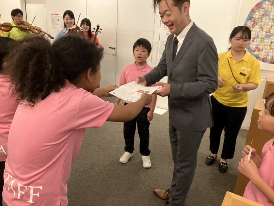 東京交響楽団の楽団長に受付でプログラムを渡すジュニア・プロデューサーの写真。周りで他のプロデューサーや、登場前にロビーで待機する出演者が笑顔で見守っている