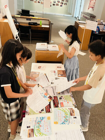会場のロビーの机にチラシ5枚程を並べて、プログラムに挟み込む作業をする広報班と運営班のメンバー