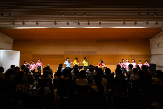 立って演奏する出演者たちと、その左右に立って並ぶジュニア・プロデューサーたちの写真