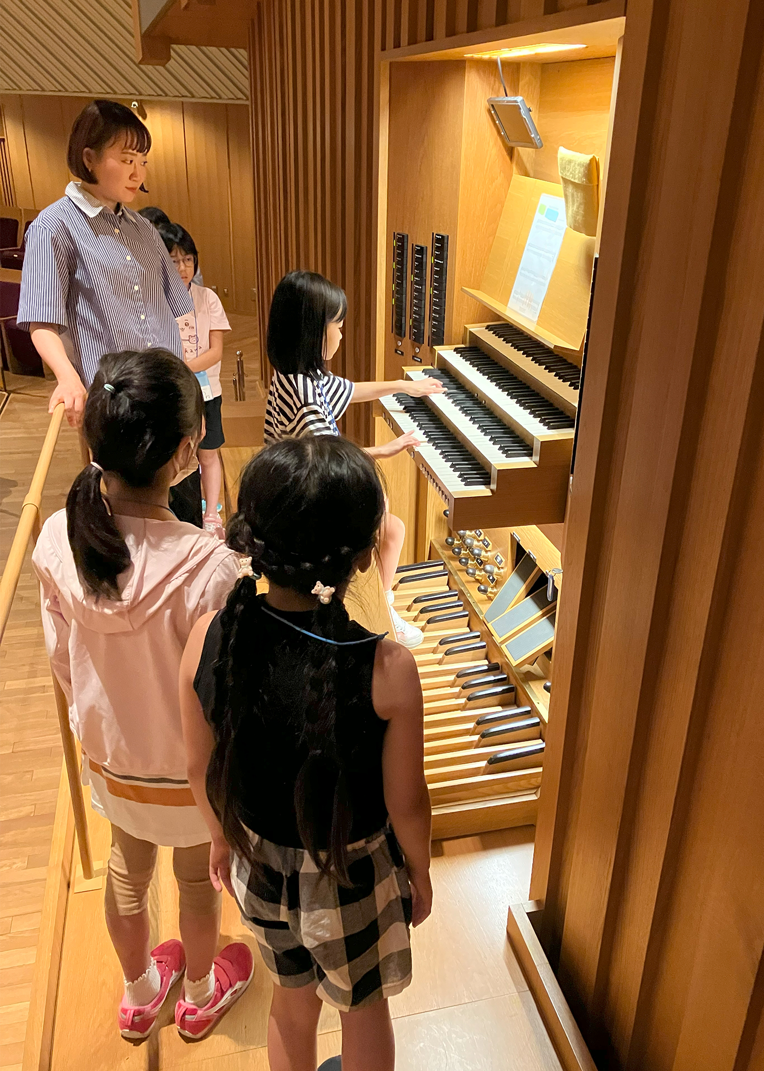 大オルガンの演奏台に座り演奏する女の子と、周りで立って見守る大木麻理さんと子供たちの写真。演奏している子は、右手を4段の手鍵盤のうち一番下の鍵盤、左手をその上の鍵盤に置いている。
