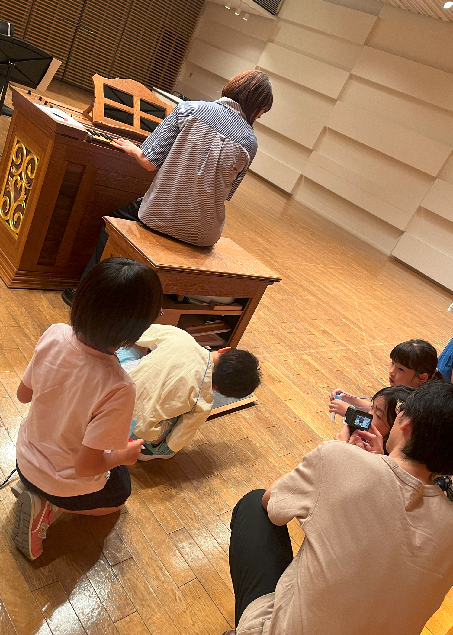 ポジティフオルガンの椅子の中にある、空気が入っている場所をしゃがんでのぞき込む子供たちの写真。大木麻理さんは椅子に座り、鍵盤に手を置きながら、後ろを振り返っている。