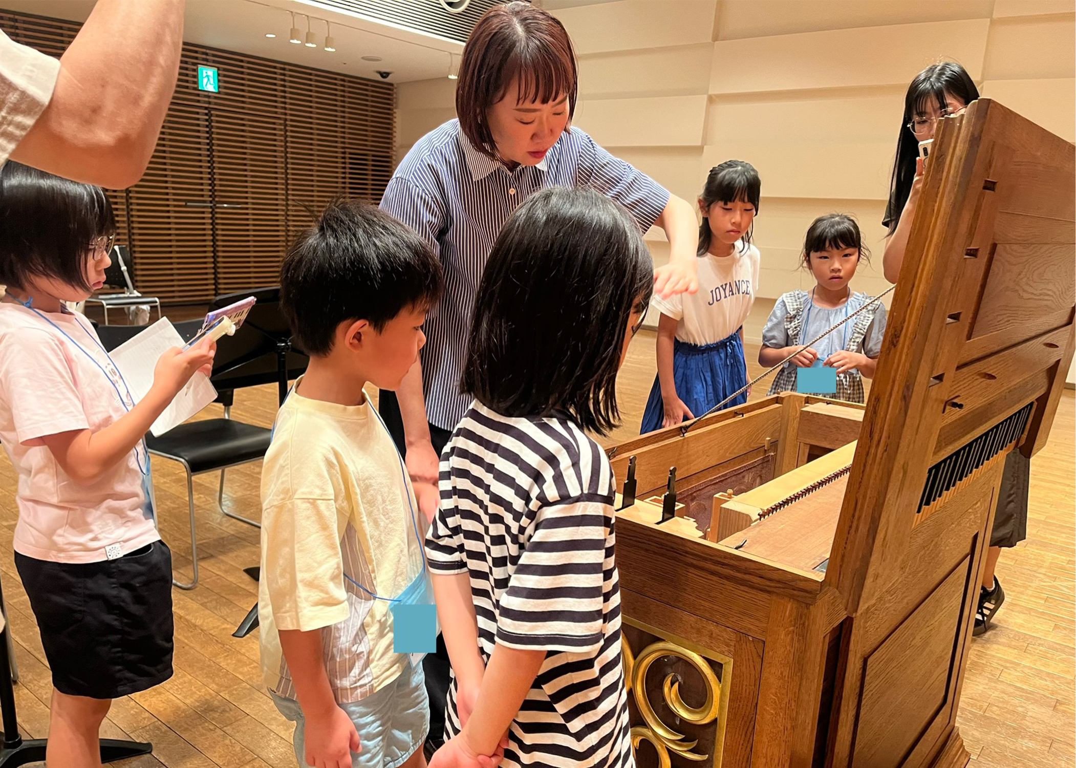 ポジティフオルガンの鍵盤を開けて、箱の中に詰まっているパイプたちを上からのぞき込む子供たちと大木麻理さんの写真。子供たちは興味深そうに前のめりになっており、その周りで保護者はカメラを向けている。