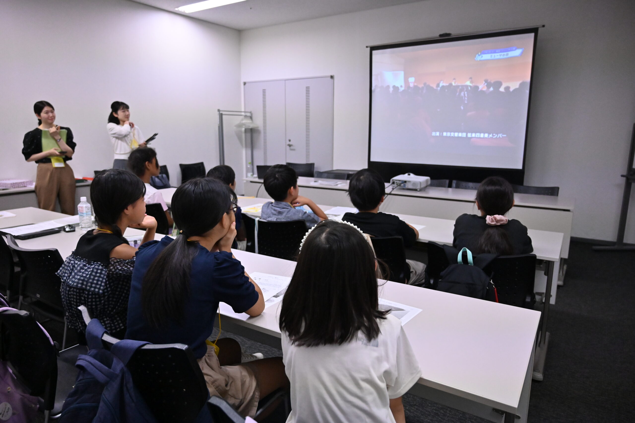 前方スクリーンに映し出されたテレビの映像を前のめりで見ているジュニア・プロデューサーたち。