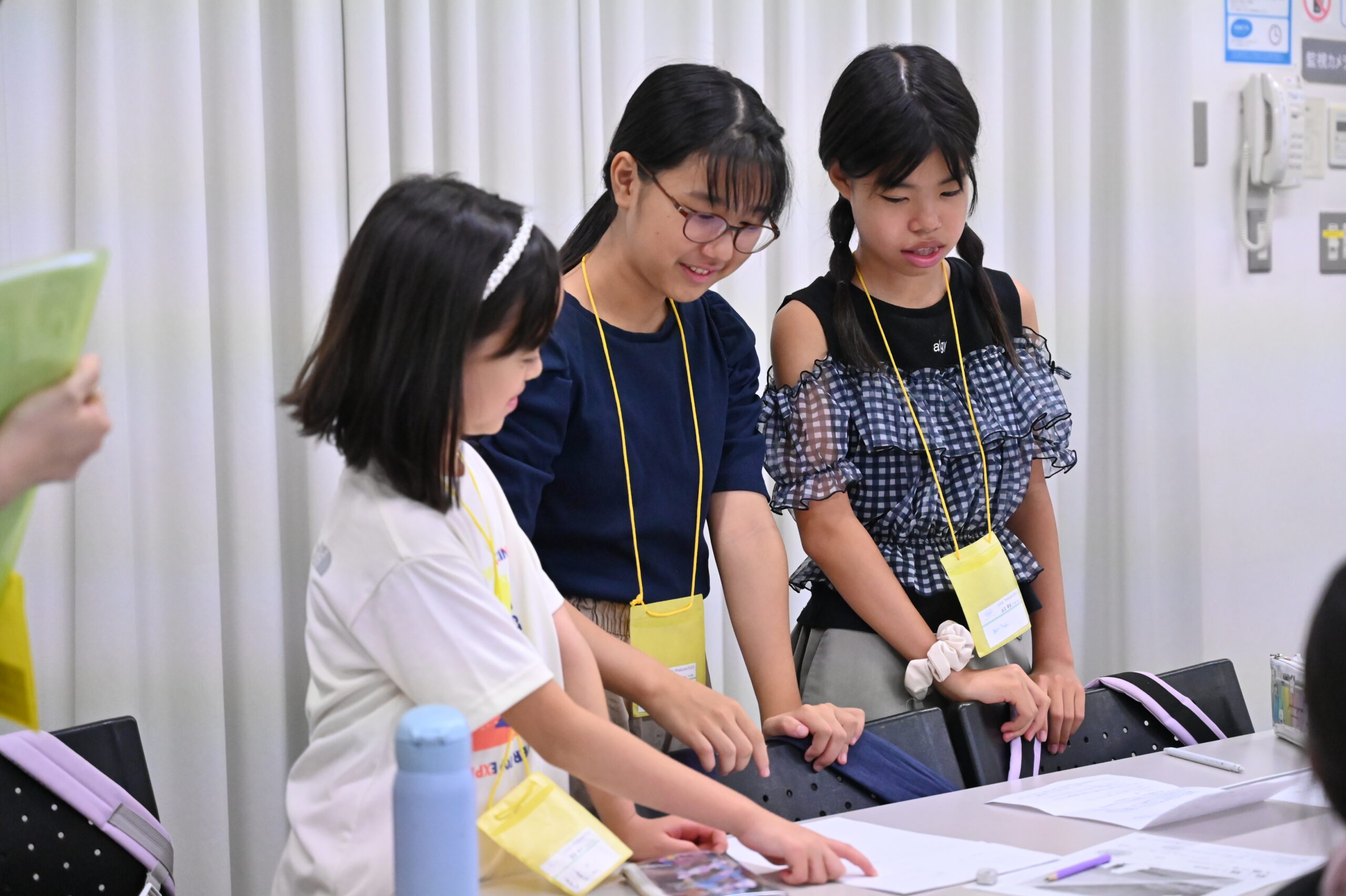 広報班3人が立って発表している様子。1人が机上のプリントを指差している。