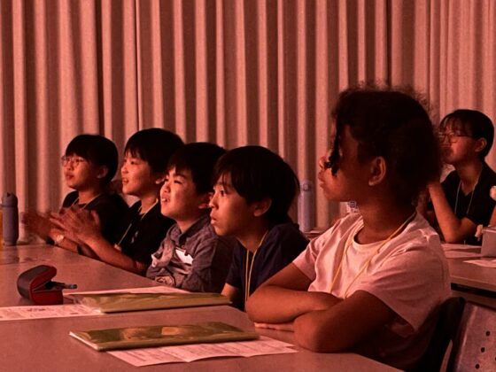 暗い部屋でコンサート映像を見ているジュニア・プロデューサーが笑顔で、拍手したりしている。