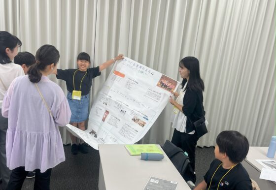 作成したジュニア・プロデューサーが両手を広げても収まりきらない大きな模造紙を大人スタッフと持ち、周りに他のメンバーや大人スタッフが興味深そうに集まっている。