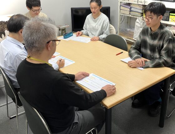 狭い打合せ室の奥にリトルミューザのメンバ2人が座り、向かいには課長と部長、横には大人スタッフが座っている。机上の各々の手元にはA4の企画書がある。