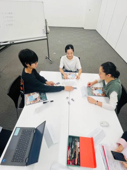 企画展示室に並べられた２台の長机に向かい合って座るリトルミューザ2025メンバー3人の写真。机上には資料やPCが広げられている。