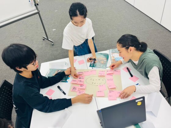 3人（学生と思われる）のグループワークの様子。大きな紙を中心に、ピンクの多数のポストイットを貼り付けながら「ミューザのイメージ」をディスカッションしている。