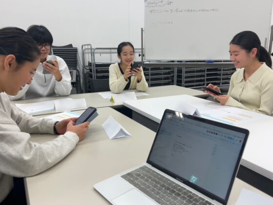 話し合いの様子。手前には開かれたノートパソコンがあり、名前プレートを前にスマートフォンを操作しながら情報収集や作業を行っている。