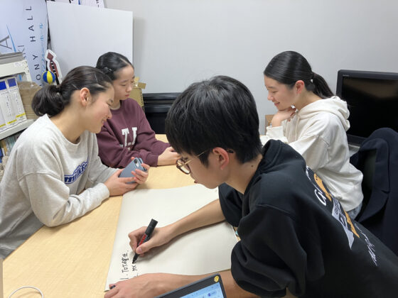 打合せ室にいる4人のメンバーの写真。奥の3人はスマホを手に話し合い、手前のメンバーが紙にメモしている。